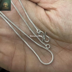 May include: A close-up of a silver chain necklace with a hook clasp. The chain has a woven design and is displayed against a skin-toned background. The necklace is a delicate piece of jewellery.