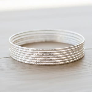 Peut inclure: Un ensemble de six bracelets argentés avec une texture martelée. Les bracelets sont ronds et présentent une surface légèrement irrégulière, reflétant la lumière. Les bracelets sont empilés sur une surface en bois de couleur claire.