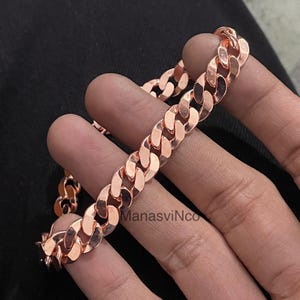 Pode incluir: Uma pulseira de corrente de meio-fio em ouro rosa. A pulseira apresenta uma série de elos ovais interligados. A joia é exibida em um fundo escuro. O nome da marca "ManasviNco" é visível.