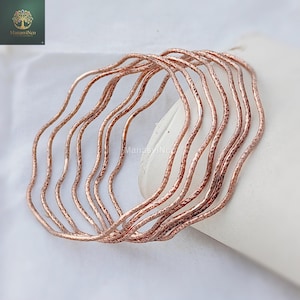 May include: A set of copper-colored bangles with a hammered texture and a wavy design. The bracelets are stacked together, creating a layered effect. The jewelry is displayed on a white surface.