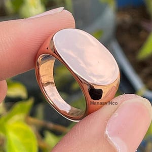 Pode incluir: Um anel de sinete em ouro rosa com uma face oval. O anel é segurado entre dois dedos contra um fundo verde desfocado. O anel tem uma superfície polida e reflexiva.