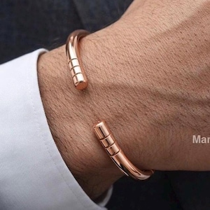 Peut inclure: Un bracelet manchette en or rose au design minimaliste. Le bracelet ouvert présente une bande lisse et incurvée avec trois lignes horizontales à chaque extrémité. Le bracelet est présenté sur le poignet d'une personne, contre une chemise blanche.