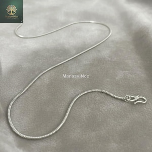 Op de afbeelding: Een zilverkleurige slangketting met een haaksluiting, gepresenteerd op een grijze stof. De ketting is dun en flexibel, met een glad, reflecterend oppervlak. De merknaam "ManasVinCo" is zichtbaar in de afbeelding.