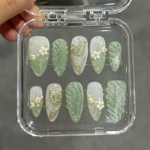 May include: A set of press-on nails in a clear plastic case. The nails feature a variety of designs, including glitter, floral accents, and gold leaf patterns. The color palette includes shades of green, white, and gold.