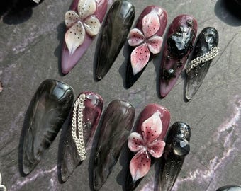 May include: A set of ten long, almond-shaped artificial nails. The nails feature a variety of designs, including marble effects, floral embellishments, and silver chain accents. Colors include black, purple, white, and pink.