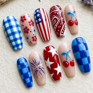 May include: A collection of press-on nails with various designs, including gingham, floral, American flag, paisley, cherries, and fireworks. A small American flag pin and a thimble are also present.