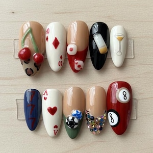 May include: A set of ten artificial nails with various designs. The designs include cherries, playing card suits, a cocktail, a billiard ball, and a soccer ball. The nails are in a variety of colors, including red, white, black, and nude.