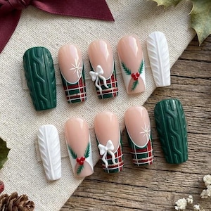 May include: A set of ten press-on nails featuring a holiday theme with dark green cable knit textures, red and green plaid patterns, white 3D bows, holly berry accents, and white cable knit designs, arranged with a deep burgundy satin ribbon.