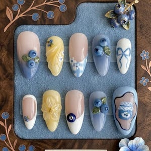May include: A set of ten artificial nails with a blueberry theme. The nails feature various designs, including blueberries, floral patterns, and a bow. Colors include blue, white, yellow, and pink. The nails are displayed in a wooden frame.