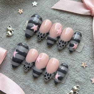May include: Set of press-on nails with a cat theme. The nails feature a combination of gray and pink colors. Some nails have gray stripes, pink stars, and cat faces. The nails are oval-shaped.