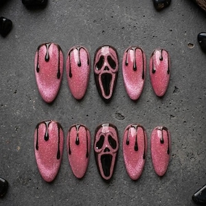 May include: A set of ten almond-shaped press-on nails featuring a shimmering pink glitter base. Each nail is decorated with a glossy black drip pattern, while two nails feature a detailed black ghost face mask design with hollow eyes and mouth.