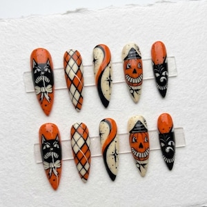 May include: A set of ten almond-shaped press-on nails featuring Halloween-themed designs in orange, black, and cream. Patterns include black cats with white bows, orange and cream argyle, black starbursts on cream, and smiling jack-o'-lantern faces.