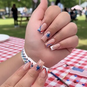 Puede incluir: Primer plano de una mano con u&ntilde;as largas y cuadradas. Las u&ntilde;as presentan una base nude con dise&ntilde;os rojos, blancos y azules, incluyendo estrellas y rayas. La manicura tiene un tema patri&oacute;tico.