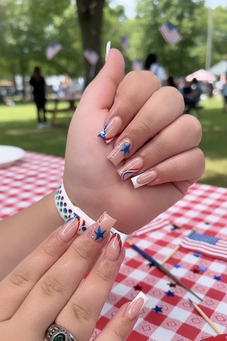 Puede incluir: Primer plano de una mano con u&ntilde;as largas y cuadradas. Las u&ntilde;as presentan una base nude con dise&ntilde;os rojos, blancos y azules, incluyendo estrellas y rayas. La manicura tiene un tema patri&oacute;tico.