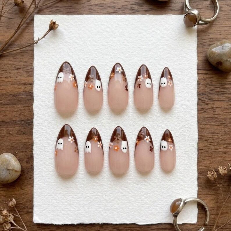May include: A set of ten almond-shaped press-on nails featuring a nude base with dark brown tips. Each nail is decorated with small white ghost figures and tiny orange and white floral accents painted across the brown tip area.