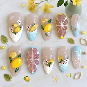 May include: A collection of ten acrylic nail designs featuring various fruit and floral themes. The designs include lemons, grapefruit, and white flowers, with accents of blue and white. The nails are arranged on a white surface with decorative elements.