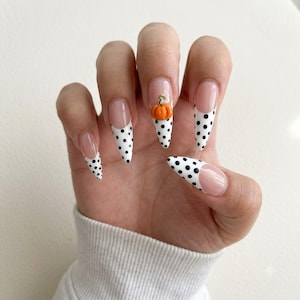 May include: Almond-shaped nails featuring a French manicure style with white tips adorned with small black polka dots. The middle fingernail includes a small, three-dimensional orange pumpkin charm with a green stem attached to the white tip area.