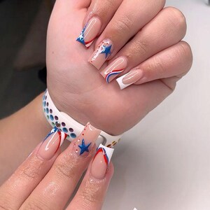 Puede incluir: Primer plano de una mano con u&ntilde;as largas y cuadradas. Las u&ntilde;as presentan una base nude con dise&ntilde;os patri&oacute;ticos, incluyendo estrellas azules, rayas rojas y azules y puntas blancas. El dise&ntilde;o recuerda a la bandera estadounidense.