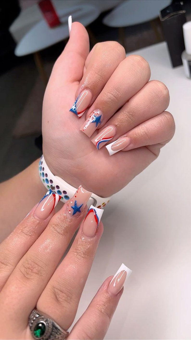 Puede incluir: Primer plano de una mano con u&ntilde;as largas y cuadradas. Las u&ntilde;as presentan una base nude con dise&ntilde;os patri&oacute;ticos, incluyendo estrellas azules, rayas rojas y azules y puntas blancas. El dise&ntilde;o recuerda a la bandera estadounidense.