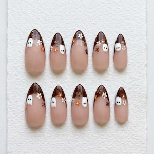 May include: A set of ten almond-shaped press-on nails with a nude base and dark brown French tips. The tips are decorated with small white ghost figures and tiny orange and white flowers. The design is consistent across all ten nail pieces.