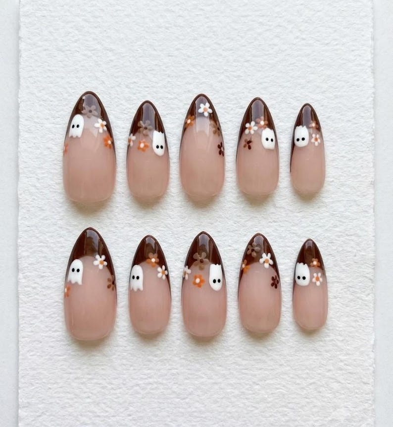 May include: A set of ten almond-shaped press-on nails with a nude base and dark brown French tips. The tips are decorated with small white ghost figures and tiny orange and white flowers. The design is consistent across all ten nail pieces.