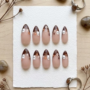 May include: A set of ten almond-shaped press-on nails featuring a nude base with dark brown French tips. Each nail is decorated with small white ghost figures and tiny brown and white floral accents scattered across the dark brown tip area.