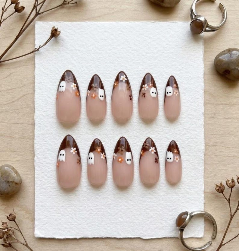 May include: A set of ten almond-shaped press-on nails featuring a nude base with dark brown French tips. Each nail is decorated with small white ghost figures and tiny brown and white floral accents scattered across the dark brown tip area.