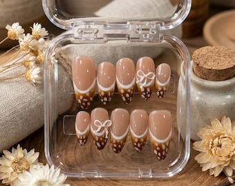 May include: A set of press-on nails in a clear plastic case. The nails are a light brown color with a white and brown dotted design. Some nails feature a white bow accent. The nails are oval-shaped.