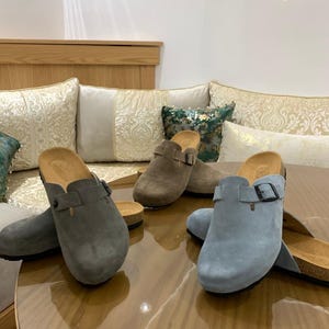 Könnte beinhalten: Eine Auswahl an Wildleder-Clogs in verschiedenen Farben, darunter Grau, Braun und Hellblau, sind auf einem glänzenden Tisch ausgestellt. Jeder Clog hat einen Schnallenverschluss und ein Korkfußbett. Die Clogs sind eine lässige Schuhoption.