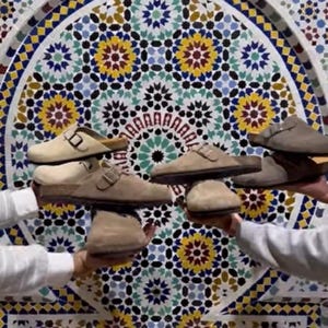 May include: Several pairs of suede slip-on shoes in various shades of beige and brown are displayed against a backdrop of a colorful mosaic tile wall. The shoes have buckle and strap details.