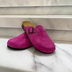 May include: A pair of bright pink suede clogs with black soles and a buckle detail. The clogs have a cork footbed and are displayed on a white marble surface.