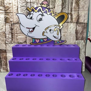 May include: A purple tiered organiser with holes, featuring a cartoon image of Mrs. Potts and Chip from Beauty and the Beast. The organiser is designed to hold pens, pencils, or small items. The background is a textured wall.