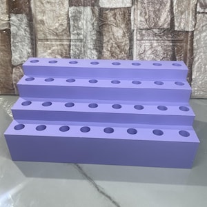 May include: A tiered, lavender-colored display stand with multiple circular holes. The stand has three levels, each with evenly spaced holes, likely for holding items. The design is simple and functional, with a smooth, matte finish.