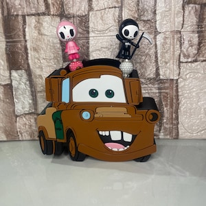 Könnte beinhalten: Ein braunes, cartoonförmiges Autozubehör mit einem lächelnden Gesicht und großen Zähnen. Zwei kleine Figuren, eine rosa und eine schwarze, sitzen obenauf. Das Auto hat grüne und beige Akzente.