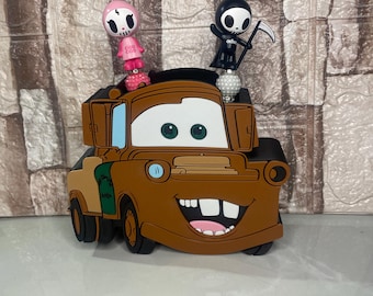 May include: A brown cartoon car-shaped accessory with a smiling face and large teeth. Two small figurines, one pink and one black, are perched on top. The car has green and tan accents.