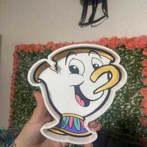 May include: A white Chip character-shaped tray with gold accents. The tray features a smiling face with a pink mouth and a blue, purple, and yellow collar. The tray is a decorative item.