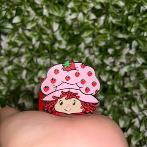 Puede incluir: Un charm rosa y rojo del personaje de Strawberry Shortcake. El charm presenta una cara sonriente, pelo rojo y un sombrero adornado con fresas. El charm se sostiene en una mano sobre un fondo de hojas verdes.