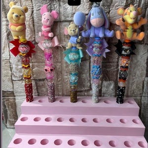 Könnte beinhalten: Fünf dekorative Stiftaufsätze mit beliebten Zeichentrickfiguren. Jeder Stiftaufsatz hat ein einzigartiges Design mit bunten Perlen, Bändern und Figuren. Der Präsentationsständer ist eine mehrstufige rosa Struktur mit mehreren Löchern.