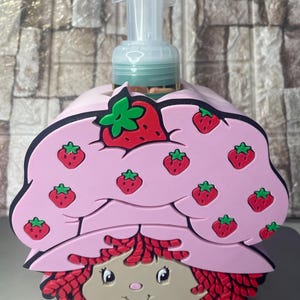 Puede incluir: Un dispensador de jabón rosa con un diseño de personaje de dibujos animados. El dispensador tiene un sombrero rosa decorado con fresas rojas y hojas verdes. El personaje tiene el pelo rojo y una cara sonriente. El dispensador tiene una bomba transparente.