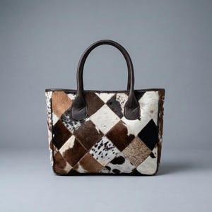 May include: A tote bag constructed from a patchwork of cowhide leather squares in shades of brown, black, and white. The bag features two dark brown leather handles attached to the top edge and a rectangular base with a structured silhouette.