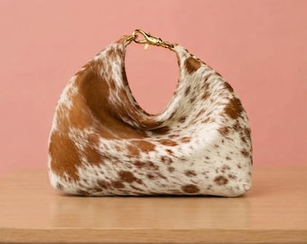 May include: A hobo-style handbag crafted from brown and white cowhide leather with a soft, textured fur finish. The bag features a distinctive curved top opening secured by a polished gold-tone metal lobster clasp hardware at the centre.