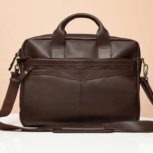 Pode incluir: Uma pasta de couro marrom com uma alça superior e uma alça de ombro removível. A bolsa tem um bolso frontal com zíper e um fecho de aba. O hardware é dourado. A bolsa está em uma superfície branca.