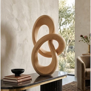 May include: A large, light-coloured wooden sculpture featuring a continuous, fluid, knotted loop design with visible wood grain patterns. The sculpture is positioned upright, showcasing a smooth, rounded surface and an open, abstract, interconnected form.