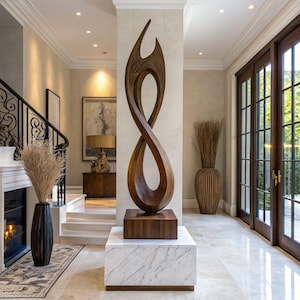 May include: A tall, dark brown wooden sculpture with a fluid, twisted, abstract form. The sculpture is mounted on a square wooden base, which sits atop a larger, rectangular white marble pedestal with subtle grey veining and a dark wood trim.