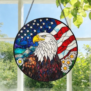 Könnte beinhalten: Runde Glasfensterkunst mit einem amerikanischen Weißkopfseeadler mit gelbem Schnabel und weißem Kopf. Der Hintergrund zeigt die amerikanische Flagge und einen blauen Himmel mit weißen Sternen. Gänseblümchen sind unten.