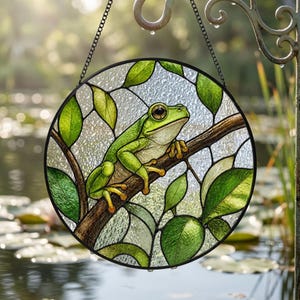 Könnte beinhalten: Ein runder Buntglas-Sonnenfänger mit einem grünen Frosch auf einem braunen Ast. Das Design umfasst leuchtend grüne Blätter und strukturierte, klare Glasscheiben in einem schwarzen Metallrahmen, aufgehängt an einer dünnen schwarzen Kette.