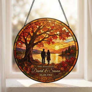 Pode incluir: Um apanhador de sol circular em vitral com uma paisagem de uma grande árvore de outono, um lago e um pôr do sol. Duas figuras estão sob a árvore. O texto diz 40th Anniversary, David & Susan e 06.08.1984. Pendurado por uma corrente preta.