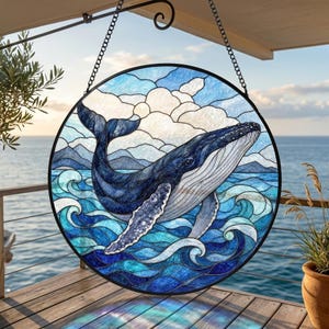 Pode incluir: Um apanhador de sol circular em vitral com uma baleia jubarte a saltar num oceano azul profundo. O design inclui nuvens brancas estilizadas, ondas ondulantes em vários tons de azul e uma moldura de metal preto suspensa por uma corrente.