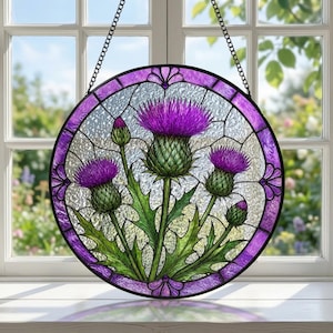 Può includere: Un acchiappasole circolare in vetro colorato con un disegno di cardi viola e verdi. L'opera è incorniciata da un bordo in metallo scuro con un anello esterno in vetro viola ed è appesa a una catena di metallo nero davanti a una finestra.