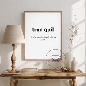 May include: A framed print displaying the word "tranquil" and its definition. It's set on a wooden table with a lamp, vases, and decorative elements. The style is minimalist and creates a sense of calm.